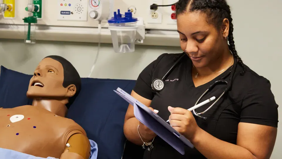 A physician assistant student wears a stethoscope and takes notes on a tablet while in a sim lab class